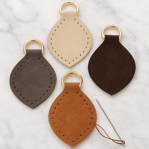 May include: Four teardrop-shaped leather bag bases in beige, brown, and tan, each with a gold-coloured ring and pre-punched holes for stitching. A needle and thread are also present, suggesting a DIY project.