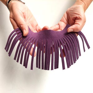 May include: A piece of purple suede leather with a fringe design. The suede is held by two hands against a white background. The fringe is made of many thin, evenly spaced strips.