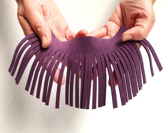 May include: A piece of purple suede leather with a fringe design. The suede is held by two hands against a white background. The fringe is made of many thin, evenly spaced strips.