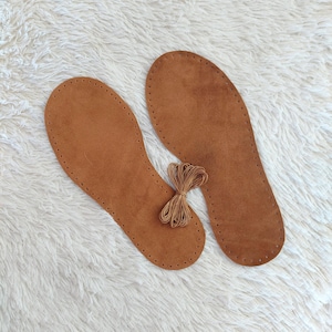 May include: Two brown leather insoles with stitched edges and a bundle of brown thread. The insoles are oval-shaped and designed to fit the inside of a shoe. The thread is tied in a small bundle.