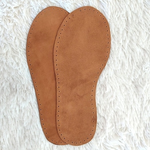 May include: Two brown leather insoles with a suede-like texture. Each insole is shaped like a foot, with a series of small holes around the perimeter for stitching. The insoles are designed for shoe making or repair.
