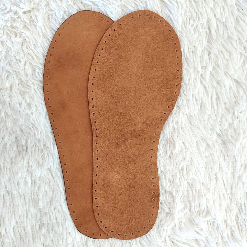 May include: Two brown leather insoles with a suede-like texture. Each insole is shaped like a foot, with a series of small holes around the perimeter for stitching. The insoles are designed for shoe making or repair.