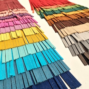 Op de afbeelding: Close-up van twee sets kleurrijke leren franjes. Eén set heeft een verloop van roze, geel, blauw en marine. De andere set heeft een verloop van rood, groen, bruin en grijs. De franjes zijn in dunne stroken gesneden.