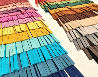 May include: Close-up of two sets of colorful leather fringe. One set features a gradient of pinks, yellows, blues, and navy. The other set has a gradient of reds, greens, browns, and grays. The fringe is cut into thin strips.