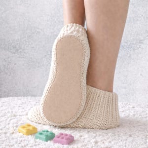 Könnte beinhalten: Cremefarbene gestrickte Hausschuhe mit hellbrauner Sohle. Die Hausschuhe sind auf einer weißen Oberfläche abgebildet. Drei bunte Spielzeugblöcke befinden sich im Vordergrund: gelb, grün und rosa.