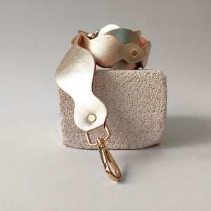 May include: A gold-colored leather wrist cuff with a floral design and a matching strap with a gold-tone clasp. The cuff is resting on a small, textured, rectangular block.