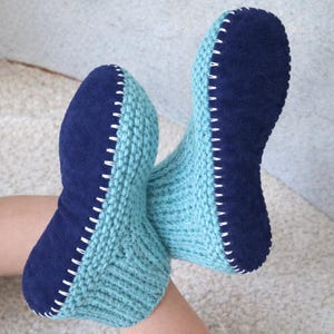 Pode incluir: Um par de pantufas feitas à mão. Apresentam uma parte superior de malha azul clara e uma sola de camurça azul escura, costuradas com fio branco. Estas pantufas são projetadas para uso interno e conforto.