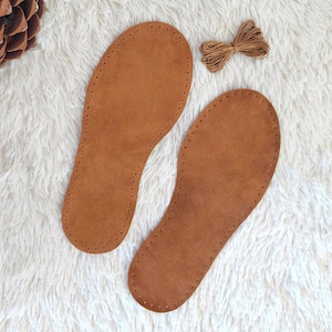May include: Two brown leather insoles with stitched edges and a bundle of brown thread on a white, fluffy surface. The insoles are shaped for the feet and have evenly spaced holes for stitching.