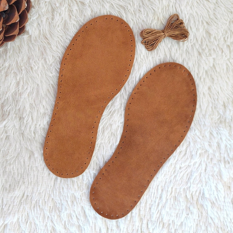May include: Two brown leather insoles with stitched edges and a bundle of brown thread on a white, fluffy surface. The insoles are shaped for the feet and have evenly spaced holes for stitching.