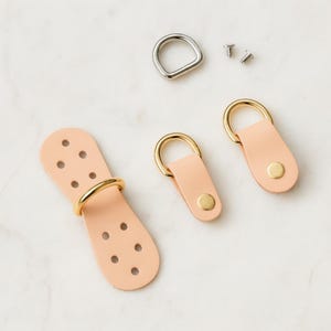 May include: A collection of leather hardware components, including two small leather straps with gold-colored D-rings and rivets, a larger leather piece with multiple holes and a gold ring, a silver D-ring, and two small screws. The leather is a light peach color.