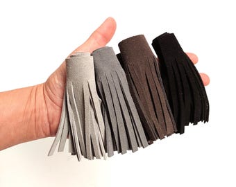 May include: Four suede tassels in shades of gray, brown, and black. Each tassel is cylindrical at the top with long, fringed edges. The tassels are held in a hand against a white background.