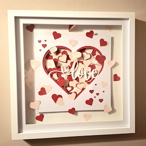 May include: A white framed artwork with a layered heart design. The heart is filled with red and pale pink hearts, with the word "love" in white script. Additional heart cutouts are scattered around the frame.
