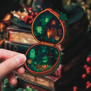 May include: A Halloween-themed sticker shaped like a pumpkin, depicting a miniature haunted house scene with ghosts, a black cat, and a witch's hat. The sticker has a vibrant, colourful design with a glowing effect.