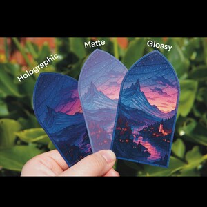 May include: Three stickers with a landscape design are held in a hand. The stickers are labeled "Holographic", "Matte", and "Glossy". The landscape features mountains, a river, and a town with a sunset sky.