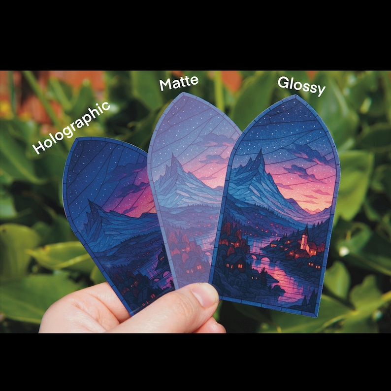 May include: Three stickers with a landscape design are held in a hand. The stickers are labeled "Holographic", "Matte", and "Glossy". The landscape features mountains, a river, and a town with a sunset sky.
