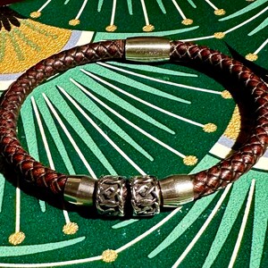 Può includere: Un braccialetto in pelle marrone con dettagli in metallo argentato. Il braccialetto ha un design in pelle intrecciata e due ciondoli in metallo decorativi.