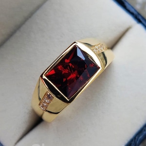 Può includere: Un anello d'oro con una grande pietra rossa rettangolare e pietre trasparenti più piccole sulla fascia.