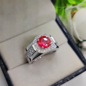 Pode incluir: Um anel de prata com uma grande pedra preciosa vermelha brilhante e redonda no centro, rodeada por pedras claras menores. O anel tem um design retangular na banda, também cravejado com pedras claras. O anel é exibido em uma caixa de joias azul escura.