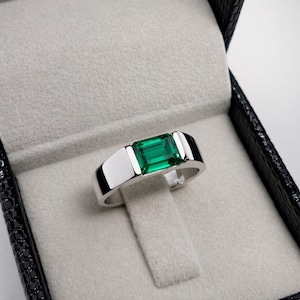 May include: A silver ring with a rectangular, emerald-green gemstone. The ring is displayed in a black jewelry box with a gray interior. The ring's band has a flat, modern design.
