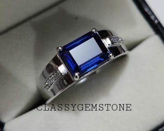 May include: A silver ring with a large, rectangular, dark blue gemstone and smaller, clear gemstones on the band. The ring is in a white box with the text "CLASSYGEMSTONE" on the bottom.