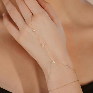 May include: A delicate gold hand chain with multiple small, clear gemstones. The jewelry piece connects to a bracelet with a thin chain. The model is wearing a black top.