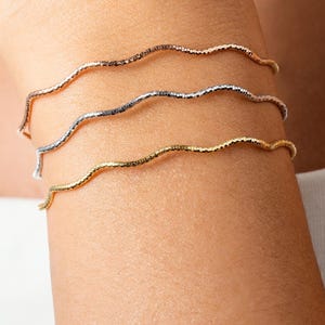 Può includere: Tre delicati braccialetti ondulati in oro rosa, argento e oro sono impilati su un polso. I braccialetti hanno un design strutturato e increspato. Il marchio HELENANDGRACE è stampato in bianco in basso nell'immagine.
