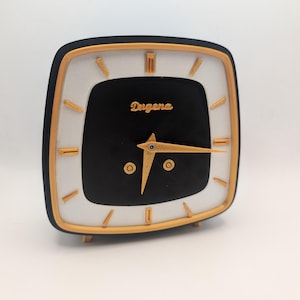 May include: A vintage square clock with a black face and gold hands, featuring the brand name "Dugena" in gold script. The clock has a white and gold border and gold accents, with small gold feet.