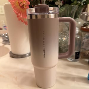 Puede incluir: Un vaso Stanley + Starbucks con asa, pajita y tapa. El vaso tiene un degradado de color, que va del beige claro en la base al beige m&aacute;s oscuro en la parte superior. El asa y la tapa son de color malva apagado.
