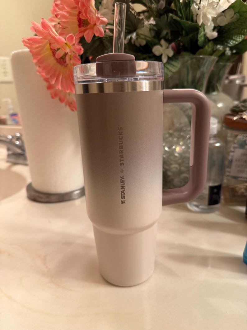 Puede incluir: Un vaso Stanley + Starbucks con asa, pajita y tapa. El vaso tiene un degradado de color, que va del beige claro en la base al beige m&aacute;s oscuro en la parte superior. El asa y la tapa son de color malva apagado.
