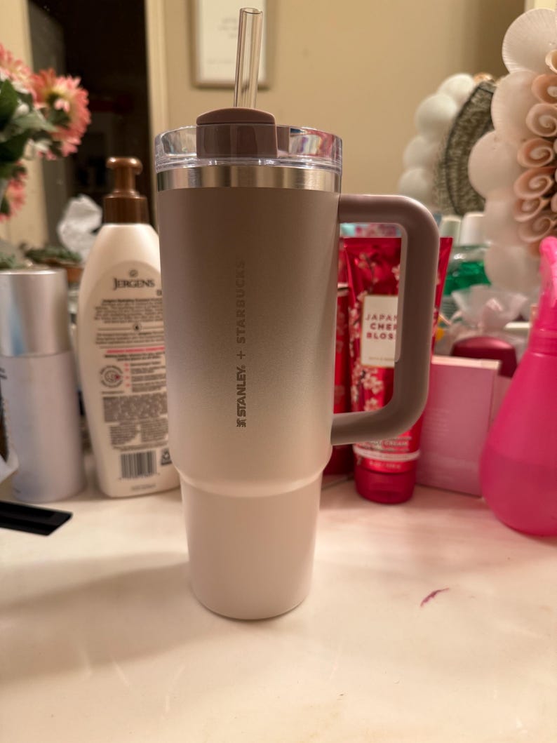 Puede incluir: Un vaso t&eacute;rmico Stanley + Starbucks con asa. El vaso tiene un degradado de color, que va del beige claro en la base al marr&oacute;n m&aacute;s oscuro en la parte superior. Incluye una pajita transparente y una tapa marr&oacute;n.