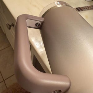 Puede incluir: Un vaso aislante de color beige claro con un asa a juego. El asa est&aacute; sujeta con herrajes plateados. El vaso tiene una tapa de pl&aacute;stico transparente y est&aacute; dise&ntilde;ado para mantener las bebidas calientes o fr&iacute;as. El vaso tiene forma cil&iacute;ndrica.