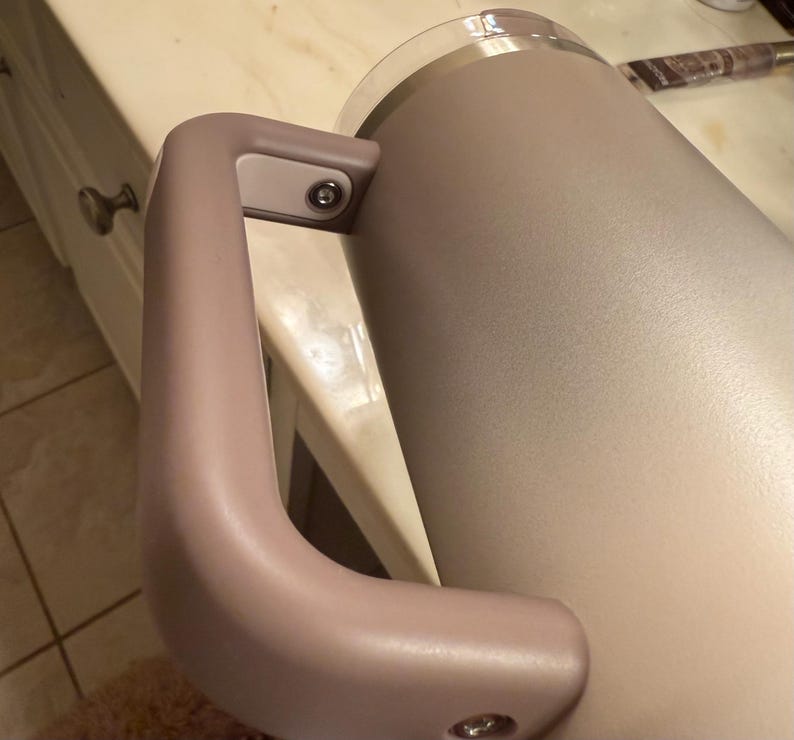 Puede incluir: Un vaso aislante de color beige claro con un asa a juego. El asa est&aacute; sujeta con herrajes plateados. El vaso tiene una tapa de pl&aacute;stico transparente y est&aacute; dise&ntilde;ado para mantener las bebidas calientes o fr&iacute;as. El vaso tiene forma cil&iacute;ndrica.