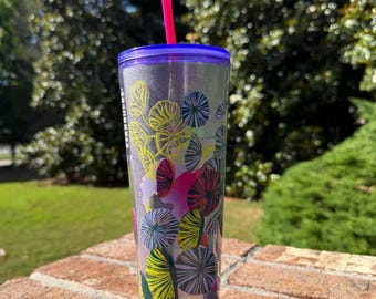 May include: A clear tumbler with a purple lid and a pink straw. The tumbler is decorated with colorful, abstract floral designs in yellow, pink, and green. The tumbler is sitting on a brick surface.