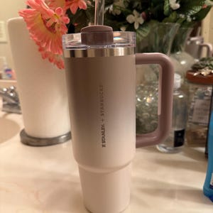 Puede incluir: Un vaso Stanley + Starbucks con asa, pajita y tapa. El vaso tiene un esquema de color degradado, que va del blanco a un marr&oacute;n claro. El asa y la tapa son de color malva apagado.