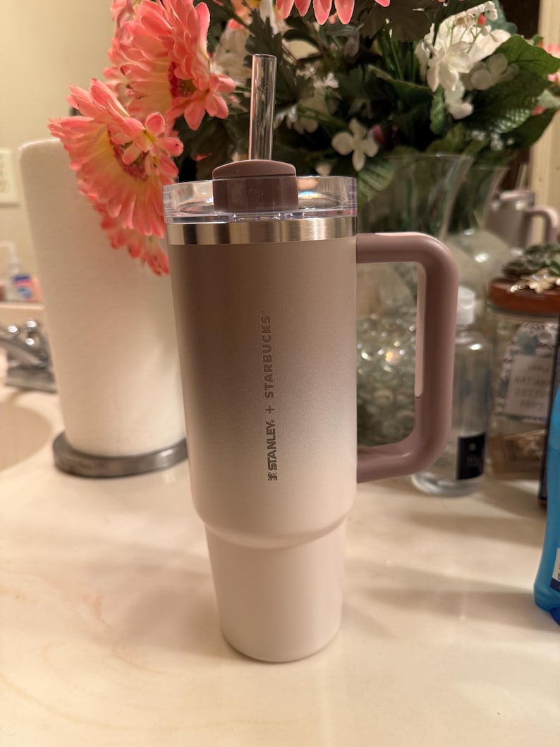 Puede incluir: Un vaso Stanley + Starbucks con asa, pajita y tapa. El vaso tiene un esquema de color degradado, que va del blanco a un marr&oacute;n claro. El asa y la tapa son de color malva apagado.