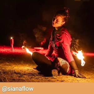 Puede incluir: Una persona con un atuendo rojo está sentada con las piernas cruzadas, sosteniendo una antorcha en llamas. La escena está tenuemente iluminada, con una segunda llama en el fondo. La persona lleva leggings negros con un diseño de red.