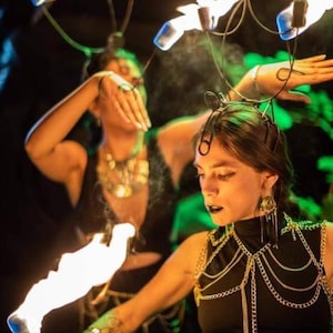 Op de afbeelding: Twee vrouwen in zwarte kleding en sieraden voeren een vuurdans uit. Een vrouw draagt ​​een hoofdband met lampjes en houdt een brandende fakkel vast. De andere vrouw houdt een brandende fakkel in haar rechterhand.
