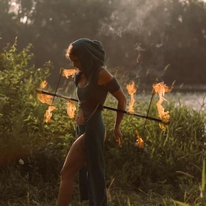 May include: A person in a teal hooded dress holding a fire staff with flames. The person is standing in a grassy area near a body of water. The image is taken during the day, with sunlight illuminating the scene.