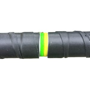 Op de afbeelding: Close-up van een zwarte tennisracketgrip met een geperforeerd oppervlak. De grip is om het handvat gewikkeld, met een gedeelte van groene en gele tape zichtbaar in het midden. De achtergrond is effen wit.