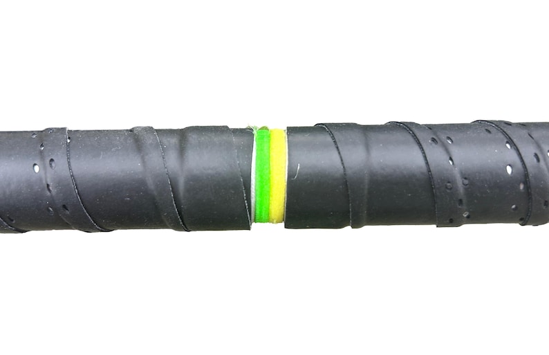 Op de afbeelding: Close-up van een zwarte tennisracketgrip met een geperforeerd oppervlak. De grip is om het handvat gewikkeld, met een gedeelte van groene en gele tape zichtbaar in het midden. De achtergrond is effen wit.