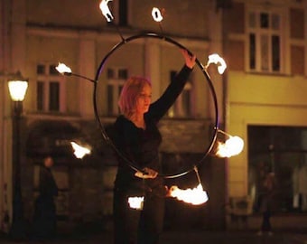 May include: A woman with pink hair performs a fire hoop routine in a city street at night. She is wearing a black shirt and black pants. The hoop is lit with fire on the ends.