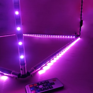 May include: A set of purple LED strip lights arranged in a triangular shape, with a remote control. The lights are mounted on silver metal tubes. The remote control has multiple colored buttons. The background is a solid purple color.