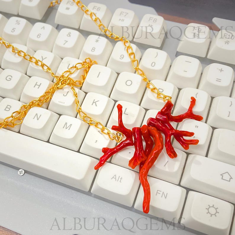 Puede incluir: Un collar de cadena de metal dorado con un colgante formado por varios segmentos ramificados de color rojo brillante que parecen coral. El collar est&aacute; sobre un teclado de computadora blanco con letras negras. El texto ALBURAQGEMS es visible.