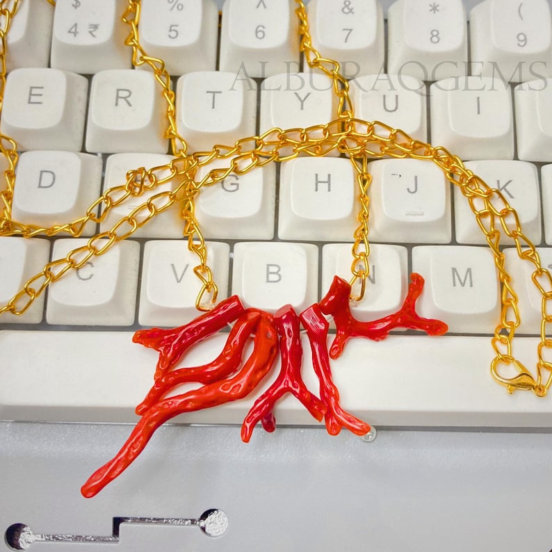 Puede incluir: Un collar con un colgante de coral rojo brillante en forma de ramas, suspendido de una cadena de metal dorado. El colgante consta de varios segmentos de coral org&aacute;nico interconectados. El texto ALBURAQGEMS es visible al fondo.