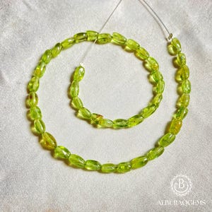 Puede incluir: Un collar de piedras preciosas de peridoto verde claro en forma ovalada. Las cuentas están ensartadas en un alambre fino y transparente, y se muestran sobre un fondo blanco. Las piedras preciosas tienen una calidad translúcida y el color general es un verde amarillento vibrante.