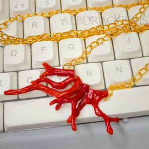 Puede incluir: Un collar con un colgante de coral rojo brillante en forma de ramas, suspendido de una fina cadena de metal dorado. El colgante consta de varios segmentos de coral rojo interconectados con formas org&aacute;nicas y una textura irregular.
