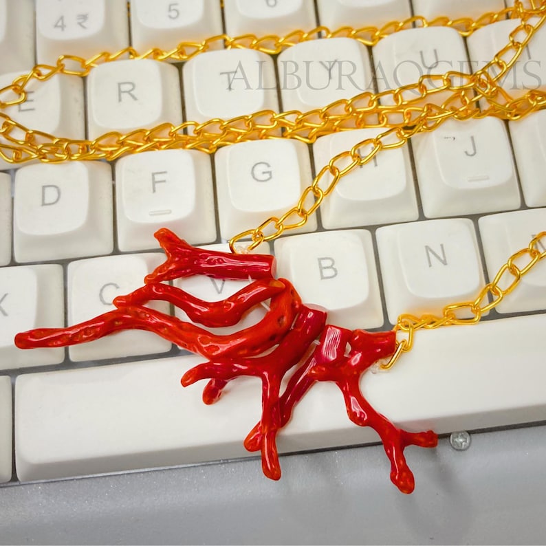 Puede incluir: Un collar con un colgante de coral rojo brillante en forma de ramas, suspendido de una fina cadena de metal dorado. El colgante consta de varios segmentos de coral rojo interconectados con formas org&aacute;nicas y una textura irregular.