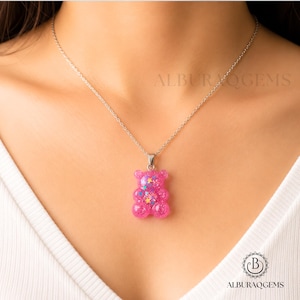 May include: A silver-toned chain necklace featuring a translucent pink gummy bear pendant. The pendant is filled with small, multi-coloured star-shaped glitter and sparkles. The text ALBURAQGEMS is visible in the background, and a logo is at the bottom.