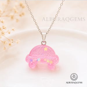 May include: A silver-toned chain necklace featuring a pink, car-shaped pendant made of resin. The pendant is filled with glitter and small, multi-coloured star-shaped sequins. The text ALBURAQGEMS is visible in the background, and a logo is at the bottom.