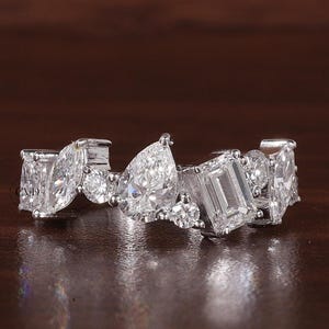 May include: A white gold ring featuring a series of diamonds in various cuts, including emerald, pear, and round shapes. The ring is set against a dark brown background, highlighting the sparkle of the gemstones.